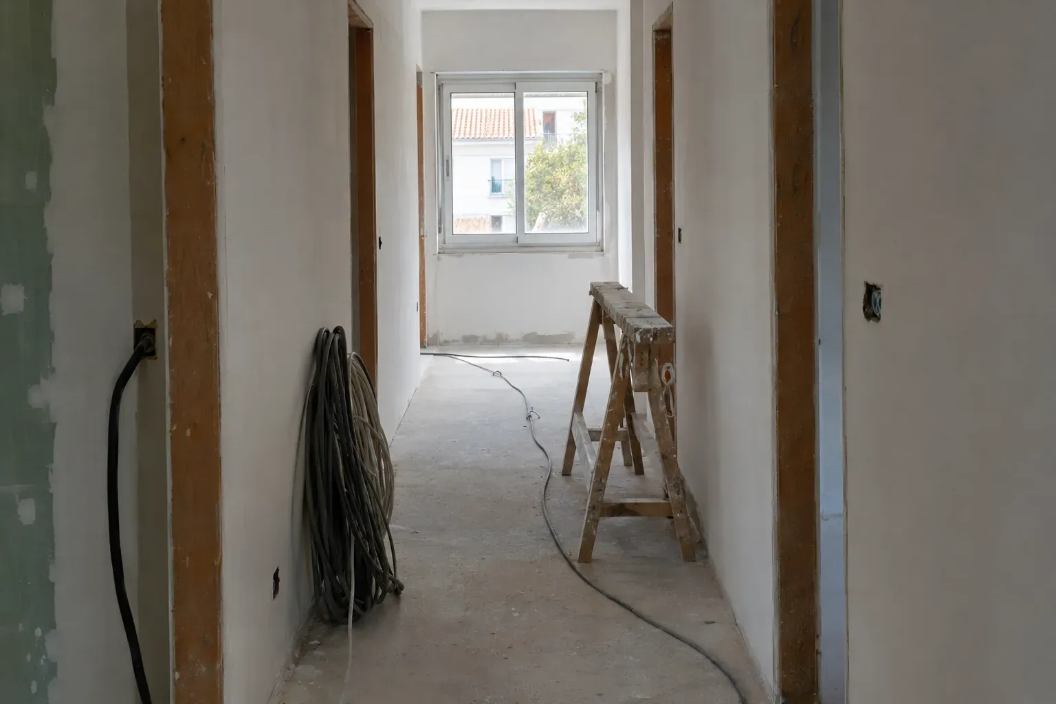 Corredor vazio de uma casa em obra, com cabos e uma janela ao fundo.