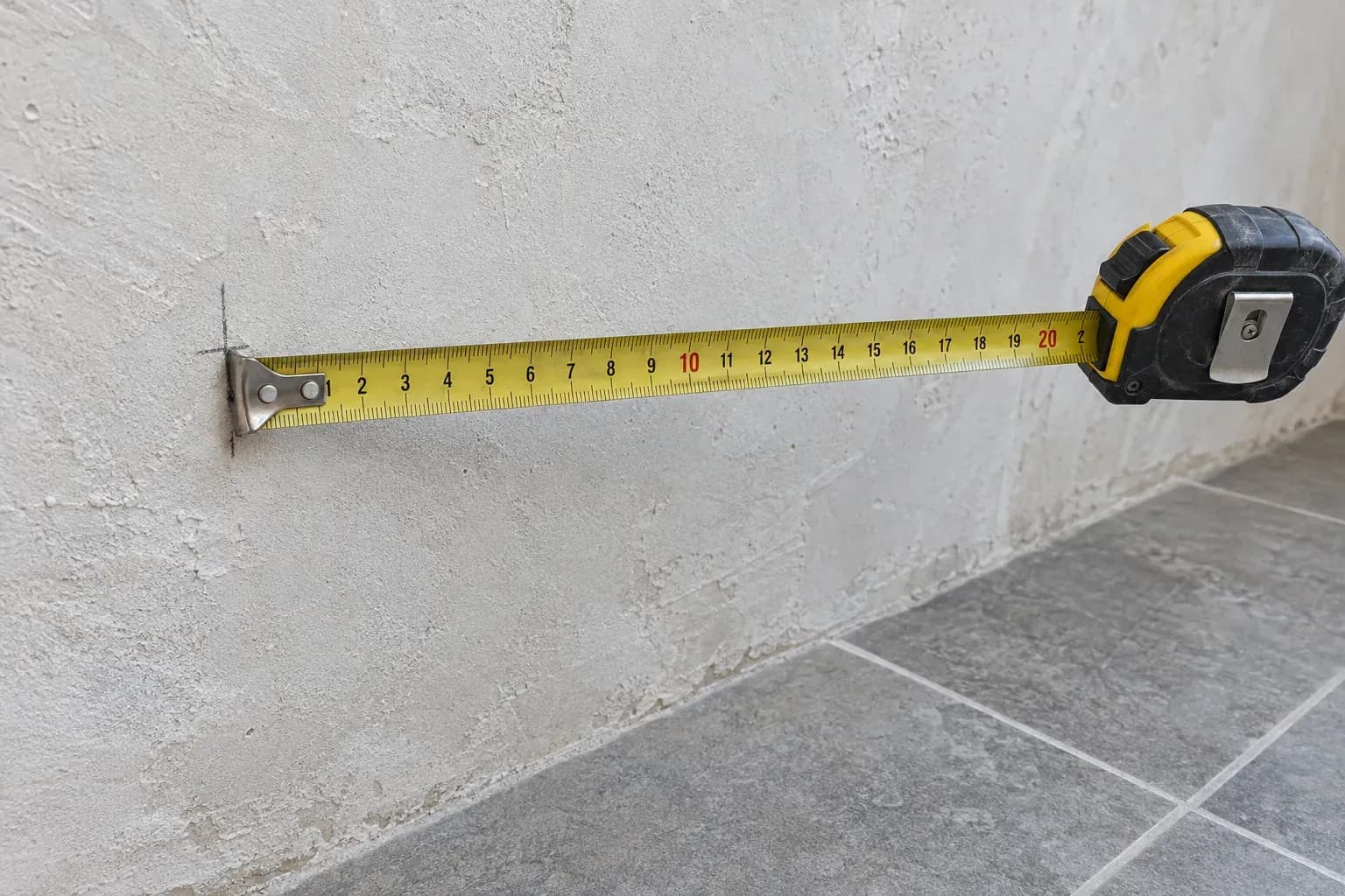 Fita métrica presa a uma parede de reboco claro, com um traço a lápis, sobre um pavimento de cerâmica.