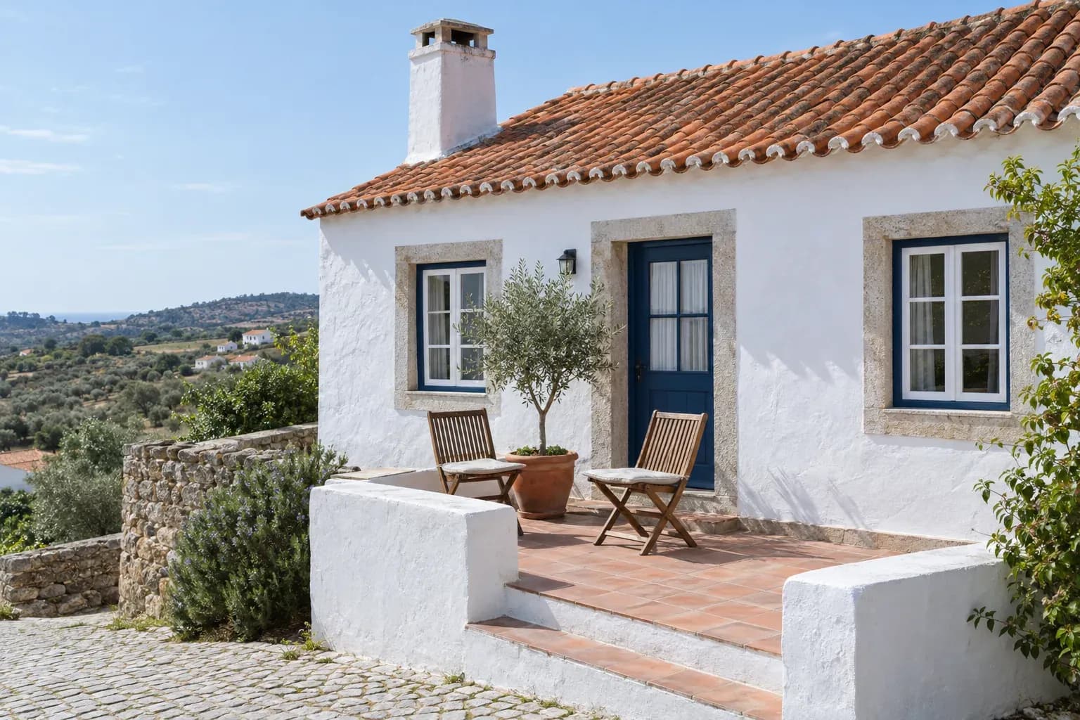 Casa caiada com telhado de telha, um pequeno terraço com duas cadeiras de madeira e uma oliveira em vaso.