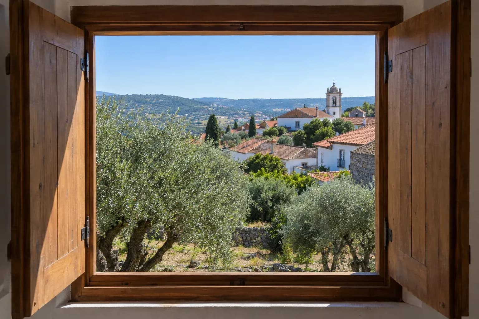 Janela de madeira aberta sobre oliveiras e casas de telhado de telha numa aldeia.